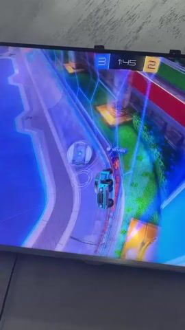 The video shows a television screen displaying the video game Rocket League. The game is in progress with the score at 3 to 2 and 1 minute 41 seconds left on the clock. A teal-colored car is driving on the field. Other players and details of the game environment are visible.