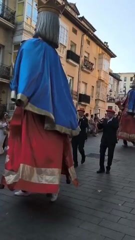 A street parade is taking place in a European town. A very large, costumed figure, appearing to be a king, with a blue cape and red garment, walks down a cobblestone street. The figure has a crown and a grey beard. Two men in dark suits and red berets walk in front of the figure, playing wind instruments. Another man in a dark suit and red beret walks beside them, carrying a drum. A crowd of onlookers lines the street. Buildings with balconies line the street, and the sky is visible above.