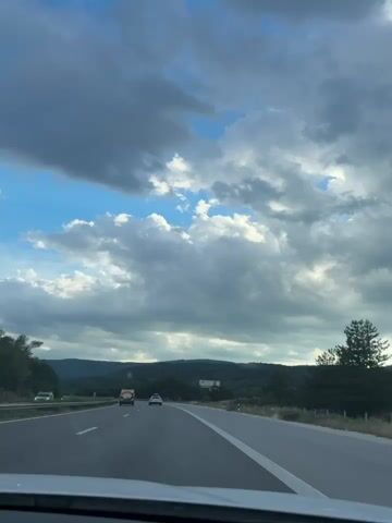 The media file shows a view from a car driving on a highway in Dolno Varshilo, Bulgaria. The sky is filled with dramatic, dark clouds interspersed with patches of bright blue sky and fluffy white clouds, suggesting a dynamic weather system, possibly before or after a storm. The sun is not directly visible, but the light suggests it is daytime, likely late afternoon given the cloud formations and lighting.

The highway is paved and has clear white lane markings, indicating it is a well-maintained road. There are several vehicles on the road, including a black SUV in the foreground on the left lane and other cars further ahead. The setting is rural, with rolling hills covered in dense green forests forming the background. The trees are lush and green, typical of a summer season.

On the right side of the road, a billboard is visible. It features a photograph of a man with a beard, possibly an advertisement or public service announcement. The logo of a circular icon with an abstract design is also visible on the billboard. The overall mood of the scene is one of travel and transit, with the natural beauty of the Bulgarian landscape dominating the view, juxtaposed against the man-made highway and advertising. The weather conditions add a sense of grandeur and perhaps a touch of mystery to the journey.