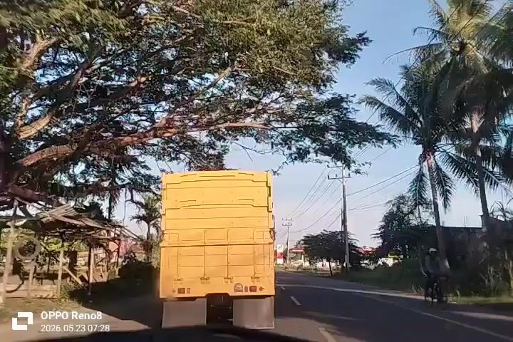 The video is shot from inside a moving car, following a large, light brown truck on a paved road. The truck has a large, boxy cargo area constructed with wooden planks. On either side of the road, there are trees, utility poles with many wires, and some buildings, including a gas station in the distance. A motorcycle passes by on the right side of the road. The sky is light blue with some clouds. The video is taken with an OPPO Reno8, and the date stamp on the screen reads 2026.05.23 07:28.