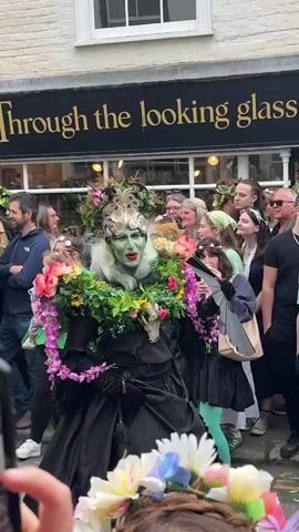 A parade of people wearing floral decorations and green face paint move past the camera on a city street. The first person in the video wears a black dress, black gloves, a headdress, and carries a black fan. The person's face is painted green, and they wear a necklace made of flowers and greenery. The group is walking past a shop named "Through the looking glass". Spectators stand along the sides of the street, some holding up their phones to record the procession.