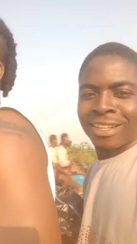 The video shows two men riding on a motorbike. One man is wearing a white tank top and has dreadlocks in a half-up style. He also has a tattoo on his left arm. The other man is wearing a gray t-shirt and is holding a black plastic bag. He turns toward the camera several times, appearing to speak. The background shows a landscape with grass and trees under a blue sky.