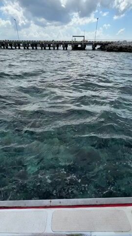 This video shows a view from a boat moving through clear, turquoise water. The water is shallow enough to see the sandy bottom and some coral formations. In the distance, a long pier extends out into the water, with a building at its end. The sky is partly cloudy with a bright blue sky visible. The boat's white deck and red trim are visible in the foreground.