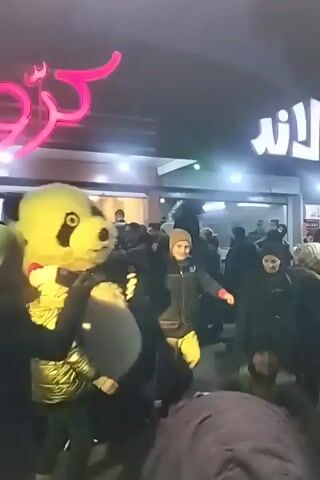 A crowd of people stand outside a building with a neon sign written in Arabic script and the words “FAST FOOD” written inside the shape of a hand making an OK symbol. A person in a panda costume with blue lighting dances in front of the crowd as people take pictures of them. The lighting on the costume changes to red.