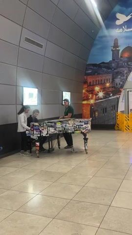 A young woman and a young man are playing a piano together. A man stands behind the piano watching them. The piano is decorated with stickers. They are in a large, open space with a tiled floor and a curved wall behind them. There are two illuminated screens on the wall. To the right of the scene is a large mural depicting a city skyline.