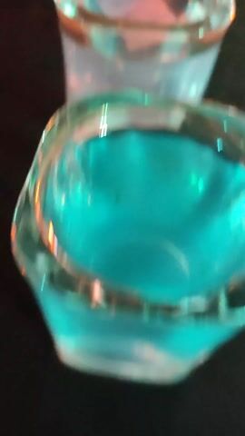 The video shows a row of small, clear glasses filled with a bright blue liquid. The glasses are arranged tightly together on a dark surface.  Confetti or similar small, colorful particles are scattered on the surface behind the glasses.  The camera angle is low, looking up at the row of glasses. The glasses appear to be shot glasses.