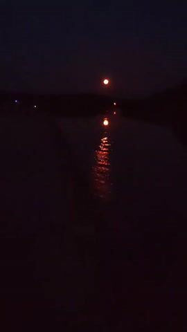 The video is a night-time shot of a body of water. The water reflects two orange lights in the distance. One light is higher and more distant, while the other is closer and appears to be on the water's surface. The sky is dark and there are faint lights visible on the left horizon. The overall mood is dark and somewhat mysterious.