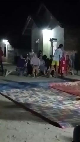 A group of people, including women and children, are gathered outdoors at night. A young man in a light blue t-shirt and yellow shorts is standing in the center of the scene, facing away from the camera. He appears to be interacting with a woman in a red and white patterned dress who is also facing away from the camera. Other individuals are seated or standing around them. Two large, rolled-up mats or carpets are visible on the ground in the foreground. A simple house with a light on is in the background. The ground appears to be dirt or sand. The overall atmosphere suggests a casual outdoor gathering or event.