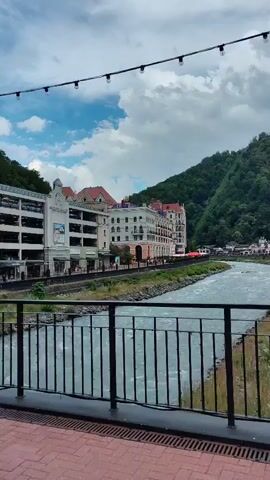 A video begins with a view of a river flowing past buildings and a mountain. A man on an electric scooter rides across a bridge. The scooter has a black frame and orange accents. The man wears a backpack. The video pans to show more of the river and the forested mountainside.