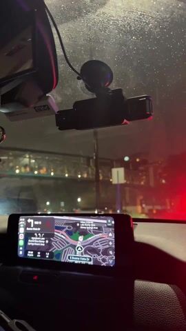 A view from inside a car, likely on a rainy night, shows a navigation system indicating the direction to Buena Vista Drive. A dashcam is mounted on the windshield. Traffic lights can be seen ahead. The dashboard is visible, as is the rearview mirror.
