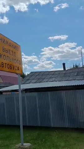 The video shows a view from a moving vehicle. The scene features a bright blue sky with scattered white clouds. In the foreground, a corrugated metal fence runs horizontally across the bottom of the frame. Behind the fence, there are several buildings, including a brick garage with a blue door, and a house with a red roof. Trees and dense green foliage are visible behind the buildings. Further back, a road sign is visible. The overall environment appears to be a rural or suburban area.