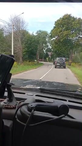 A first-person perspective video shows a car driving down a two-lane road. The road is lined with trees and vegetation. There are several other vehicles on the road, including a white car and two dark-colored SUVs. The white car is approaching from the opposite direction. The SUVs are in front of the car filming. There are pedestrian crossing signs on the side of the road. The interior of the filming car is visible, including a phone mounted on the dashboard and some decorative items. The weather appears to be overcast.