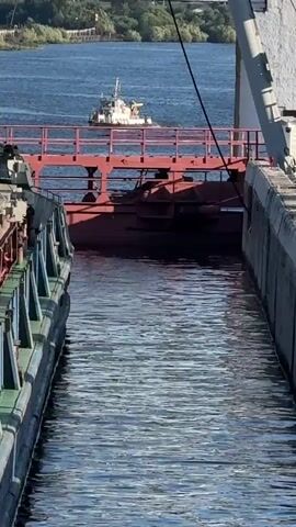 A video shows a tugboat on a body of water. The tugboat is white and has a tan superstructure. It is moving away from the camera. The water is blue and appears calm. The background is a blurry green landscape. The camera is focused on a large barge or ship, which takes up the majority of the frame. A portion of a red bridge or ramp is visible in the center of the frame, spanning across the water between two parts of the larger vessel. The sides of the large vessel are detailed with metal railings and industrial components. The environment appears to be a port or waterway, with buildings and trees visible in the distance. The sun is shining, creating reflections on the water.