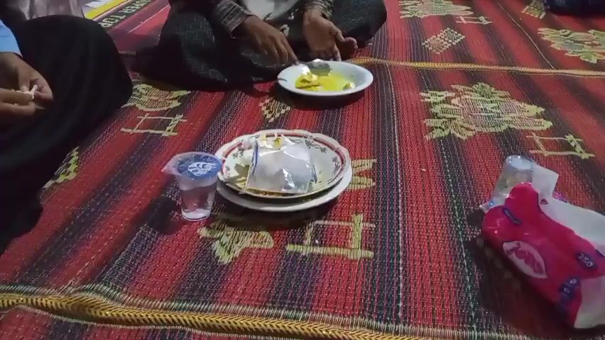 The video shows a group of people sitting on a woven mat. One person in the center is holding a small plate with food and eating from it with a utensil. In front of them are two stacked plates, one of which has a plastic covering, and a plastic cup with a blue lid. To the right of the plates is a packet of tissues. Another person is partially visible on the left, holding a white object, possibly a tissue, in their hand. A third person is visible in the background, seated further away. The environment appears to be indoors, with a black fan visible in the background.
