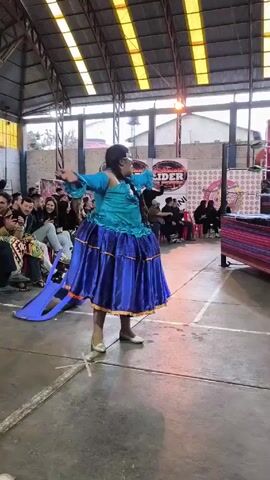 The video shows a person in a teal and blue dress dancing in what appears to be a gymnasium or indoor event space. The person is facing away from the camera and twirling. A group of people is seated on chairs along one side of the dance area watching the performance. A blue plastic chair is lying on its side near the performer. Banners are visible in the background, including one that says "Lider." The floor has white lines painted on it, typical of a gym setting. Natural light is coming through the windows in the ceiling.