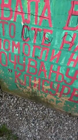 This video shows graffiti on a concrete structure, possibly a bridge or underpass. The graffiti includes large, red Cyrillic letters and some smaller white and black markings. There are also drawings of a daisy and a heart. The structure is covered in moss and dirt, and vegetation can be seen growing around it. The video pans from the graffiti-covered wall to an upward view of the concrete structure and the surrounding trees and sky.