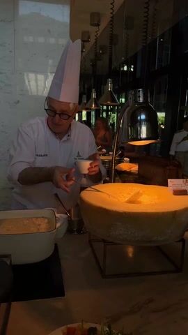 Here's a description of the video:

The video shows a chef in a white uniform and tall chef's hat, standing behind a counter in a restaurant. The chef is wearing glasses and has a bandage on his forehead. He is holding a small white bowl in his hands and appears to be preparing or adjusting food from one of two white pots in front of him. On the right side of the frame is a large wheel of cheese. Above the cheese wheel is a heat lamp. Behind the chef, partially visible in the mirrored background, are other people and food service stations. The environment suggests a high-end breakfast or brunch buffet setting. The chef's uniform has an embroidered logo on the left breast. The environment is well-lit with hanging lamps.