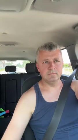 A man with short gray hair, wearing a dark blue tank top and a seatbelt, is sitting in the driver's seat of a car. He is looking forward and to the right. The interior of the car is visible, including the headliner, dashboard, and the back seats. Outside the car, trees and a building are visible. The car appears to be moving.