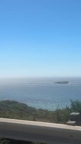 The video shows a view of the ocean from a moving vehicle. The ocean is blue and appears to be choppy. There is a small island in the distance. The foreground is partially obscured by a metal guardrail. The vegetation on the coastline is green and bushy. The sky is clear and blue.