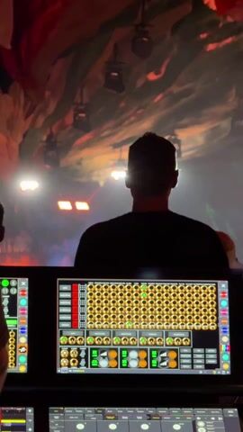 The image captures a silhouetted individual, viewed from behind, operating a control station at a live event. The person stands before a brightly lit stage or performance area, where dynamic, colorful lights cast an energetic atmosphere. The background features a large screen or projection creating an abstract, textured ceiling effect with hues of red, blue, and purple, illuminated by numerous spotlights and moving light fixtures.

In the foreground, a detailed digital control screen is prominently displayed on a console. The screen shows a grid of circular buttons and indicators, largely gold and green, alongside various sliders and text labels. Visible text on this interface includes "SPOT," "MOOD FX," "SHUTTER," "BLACKOUT," "MASTER STAGE," "ALL FIXTURES," "MAIN RGB PIXELS," "DMX IN," "ARTNET IN," "DMX OUT," and "ARTNET OUT," indicating it is a professional control system for lighting or visual effects. Below the digital screen, a physical control panel features buttons labeled "PLAY," "STOP," "GO," "BACK," "PAUSE," "NEXT," "FLASH," and "CHASE." The scene suggests an indoor or nighttime event, such as a concert or festival, with the operator actively managing the visual elements of the performance. The location context of Indio, United States, implies a large-scale event.