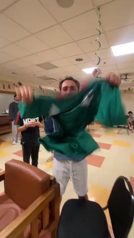 A person is holding up a green soccer jersey with red and white accents, likely the Mexican national team jersey, in front of their face and shaking it. The person is wearing a blue lanyard around their neck and white shorts. The background appears to be a classroom or common area with beige walls, a tiled floor, and fluorescent lighting. Other people can be seen in the background, and there are some decorations, including green streamers and flags. The person is blurry due to movement.