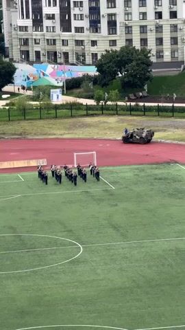 A military band marches in formation on a green sports field, with a military vehicle parked on a red track behind them. In the background, several apartment buildings and trees are visible. The scene appears to be a rehearsal or preparation for an event.