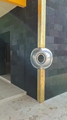 The video pans across the exterior of a building with dark grey stone tile cladding and a yellow overhang. A gold-colored decorative column with intricate carvings runs vertically up the corner of the building. Attached to the column, at waist height, is a circular, silver-colored metallic object with a hole in the center. To the left of the column, a dark doorway is visible. Further down the wall, there are large, ornate gold doors. The sun is shining brightly, casting a lens flare in the upper right of the frame. The ground in front of the building appears to be concrete. A bird is visible flying in the clear blue sky.