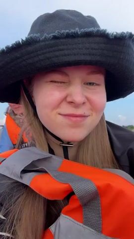 A young woman with blonde hair and a black bucket hat is on a boat. She is wearing a life jacket over a dark jacket. She winks at the camera. The sky is cloudy and there is water and land in the background.