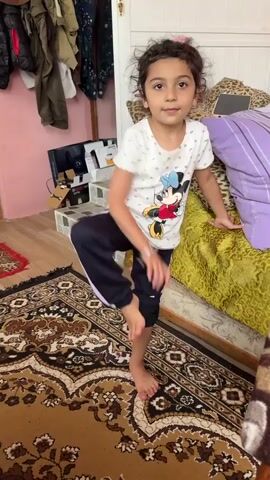 A young girl with curly dark hair and a slight smile is captured in a video performing a gymnastic split. She is wearing a white t-shirt with a Mickey Mouse graphic and dark pants. She stands on a patterned rug in what appears to be a room, possibly a bedroom. To her left, there are clothes hanging on a rack, a black box, and some other items on a tiered shelf. Behind her, a bed is visible with colorful pillows and a green textured throw. A door with a white handle is also in the background. The girl's right leg is extended straight up in the air, held by her hand, while her left leg remains on the floor, bent at the knee. The lighting suggests it is daytime. The girl's pose and expression indicate a sense of accomplishment and playful confidence. The rug's intricate pattern and the general room decor suggest a home environment, with the "Location context: Toğanalı, Azerbaijan" providing a specific geographical setting.