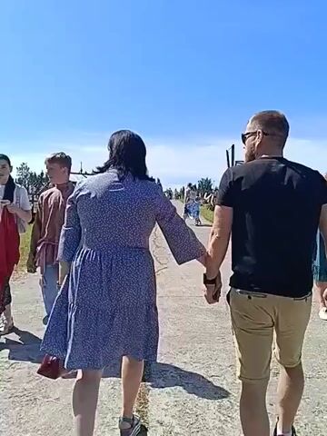 Two individuals, a woman and a man, walk away from the camera holding hands on a paved path. The woman is on the left, wearing a blue floral dress and carrying a red purse. The man is on the right, wearing a black t-shirt and tan shorts. In the background, other people are visible, along with trees, fences, and structures that suggest an outdoor event or park. The sky is clear and blue, indicating sunny weather.