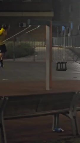 A person is swinging on a swing set at night. The person is wearing a yellow shirt and black shorts. The swing set is located in a playground, with a brick building in the background. There is a bench in the foreground, and some trash is visible on the ground near the bench. The swing set has two swings, and the person is on one of them. The person is swinging back and forth, and the swing is in motion. The scene is dimly lit, and the lighting suggests it is nighttime.
