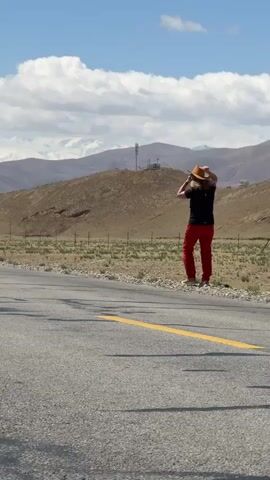 A woman is seen walking and posing on the side of an asphalt road in a vast, mountainous, and arid landscape. She is wearing a black t-shirt, red pants, and a straw hat. The road features a solid yellow line and a dashed yellow line. In the background, rolling hills with sparse vegetation lead up to distant, snow-capped mountains under a bright, clear blue sky with white clouds. A telecommunications tower or similar structure is visible on a mid-ground hill. The woman initially walks from the right side of the frame towards the left, adjusting her hat, then kneels down briefly in a dynamic pose, and stands up again. The scene is brightly lit by sunlight, casting distinct shadows on the road, indicating daytime. The overall impression is one of open space and natural beauty, characteristic of a remote location like Mainpu township, China.