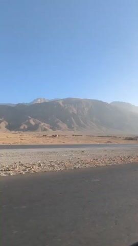 The video presents a wide, flat landscape beneath a clear blue sky. A large mountain range dominates the horizon, its slopes appearing arid and brown. In the mid-ground, a flat expanse stretches out, covered in a mix of sand and small rocks. Faint, distant structures that appear to be single-story buildings are visible. The camera appears to be moving on a paved road, with the road surface occupying the foreground. There are no people visible in the frame. The overall impression is of a remote, desert-like environment.