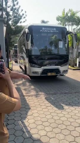 The video opens with a medium shot of a white and black passenger bus, identified as "ANB TRANS PARIWISATA," parked on a patterned paved surface. The bus has "Mercedes-Benz" branding visible on its side. A person, partially visible from behind and wearing a tan uniform, stands on the left, waving. The camera pans slightly to the right, showing the front of the bus, with people visible inside through the windshield. A license plate, "H 7396 DO," is visible on the front bumper. The bus appears to be stationary. The scene is outdoors, with trees and palm trees in the background, suggesting a warm, sunny day. The camera continues to pan right, focusing on the passenger door of the bus, where a man in a white collared shirt is seen looking out. The video ends with a close-up of the side of the bus near the door.