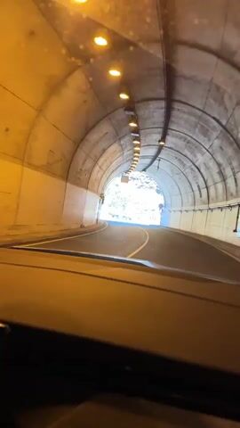 The video shows a car exiting a tunnel. The tunnel has a concrete arch shape with evenly spaced lights along the ceiling. As the car exits, the view opens up to a crossroad under a mountain. The mountain is lush with greenery and rocky outcrops. Various road signs are visible, directing traffic and indicating a pedestrian crossing. The sky above the mountain is clear blue with a few clouds. A dark colored car is parked nearby. The perspective is from inside the moving vehicle.