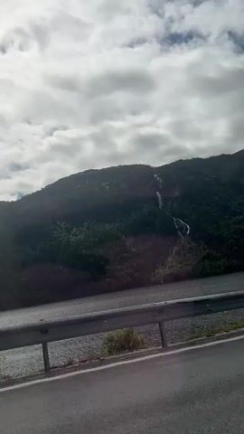 The video captures a scenic view of a mountainous landscape with a body of water in the foreground. The mountains are covered in dense, dark green vegetation, with a visible waterfall cascading down one of the slopes. The water appears dark and still, reflecting the overcast sky. In the foreground, a paved road runs alongside the water, separated by a metal guardrail. The sky is filled with dramatic, grey clouds, suggesting a cloudy or potentially rainy day. The lighting indicates it is daytime, likely late morning or afternoon given the brightness. The setting appears to be Fjæra, Norway, as suggested by the prompt, known for its dramatic fjords and natural beauty. A small, red and white flag with text and a logo is visible briefly on the right side of the road, near the guardrail, fluttering in the breeze. The overall mood is serene and majestic, highlighting the raw natural beauty of the Norwegian landscape.