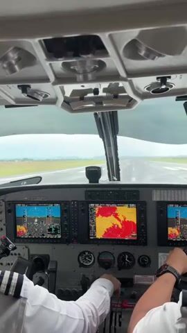 The video provides a first-person perspective from the cockpit of a small airplane. Two pilots are visible, their arms and hands on the controls. The pilot on the left wears a white shirt with striped epaulets, and the pilot on the right has their hand on the throttle. The dashboard is filled with various screens displaying flight information and a weather radar, as well as analog gauges. Outside the windshield, a runway is visible, stretching into a hazy, overcast sky. The plane appears to be on the runway, possibly preparing for takeoff or landing, indicated by the hands on the throttles and the view of the runway ahead. The overall environment suggests an active flight operation in potentially challenging weather conditions.