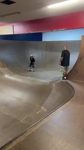 The video shows a person in a skatepark, possibly an indoor facility due to the lighting and ceiling. The skatepark has wooden ramps and curves. In the first few seconds, a man is standing near a large ramp, and a child on a skateboard is riding towards him. The man appears to be tossing something, possibly a ball, to the child. The child then rides up the ramp and performs a trick in the air, landing back on the skateboard. The video then cuts to a shot of the child standing in the skatepark with their skateboard. The environment appears to be a recreational facility designed for skateboarding.
