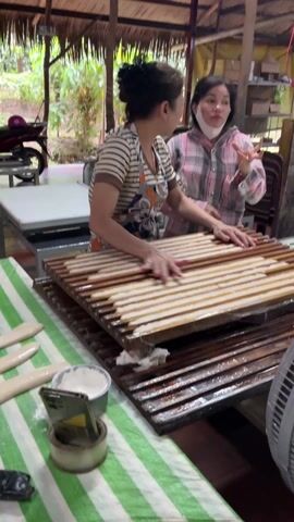 Two women are working in a rustic indoor area. One woman, wearing a striped shirt and apron, flattens pieces of dough on a grooved wooden rack. A second rack sits below it. The second woman, wearing a pink and grey plaid shirt and a face mask, observes and eats something. A bowl of flour and a smartphone are visible on a green and white striped tablecloth. A fan is running in the foreground. A motorcycle and other objects are visible in the background. The roof is thatched. The women are talking.