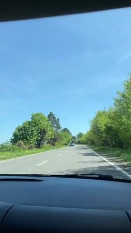 The video captures a first-person view from inside a car, looking out through the windshield on a clear, sunny day. The car's dashboard is visible in the lower portion of the frame, with the road stretching ahead. Several circular suction cups are attached to the windshield. The road is asphalt, marked with a dashed white line in the center, indicating a two-way street. Green trees and foliage line both sides of the road, creating a natural corridor. The sky above is a bright, cloudless blue. Sunlight is evident, casting shadows from the trees onto the road. In the distance, a blue car is seen driving in the opposite direction. The scene depicts a drive along a pleasant, tree-lined road. No visible text is legible in the frame.