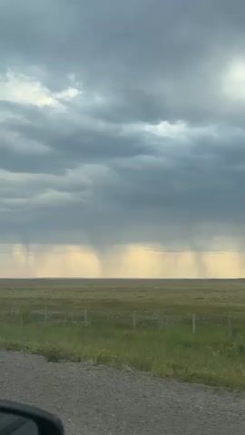 A view from inside a vehicle shows a vast, flat landscape under a dramatic, cloudy sky. Several streaks of rain are visible in the distance, falling from dark clouds onto the green and yellow field. The foreground of the image shows the edge of the vehicle's side mirror and a dirt road. The scene suggests a stormy weather event occurring in an open, rural area.