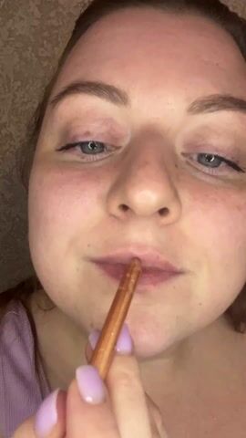 A young woman with light skin and freckles is applying lip liner to her bottom lip. She is wearing a light purple top and has pale lavender nail polish. Her eyes are blue and lined with black eyeliner. The background appears to be a textured wallpaper. The camera is close to her face, focusing on her lips and the lip liner application.