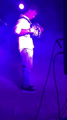 A man stands on a stage lit by purple lights, playing an accordion. He wears a white shirt and ripped jeans. To his left, a woman stands near a microphone stand. A large speaker is visible on the right side of the stage. Smoke or fog fills the air, illuminated by the stage lighting.