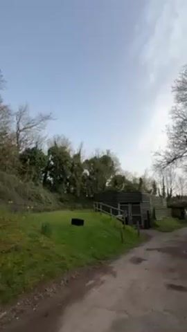 The video shows a rural scene with a grassy hill, a small green shed, and a dirt path under a partly cloudy sky. There are bare trees on the side and yellow daffodils on the hill. Someone counts down from three, and then a black box on the grass emits a large cloud of white smoke.