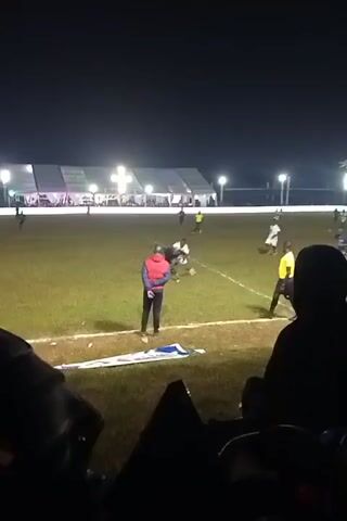 The video shows a soccer match being played at night. Players in blue and white uniforms are seen on the green field. A coach in a red vest stands near the sidelines, gesturing to the players. There are multiple bright lights illuminating the field, and a large white tent-like structure is visible in the background. Some spectators are visible in the foreground, partially obscuring the view. The game appears to be in progress, with players passing and moving the soccer ball across the field.