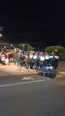 An outdoor concert is taking place at night. A band of musicians is seated on green chairs, playing various instruments including brass and woodwinds. A conductor stands in front of them, leading the performance. There are musical stands with sheet music, speakers, and audio equipment set up. The background shows trees and a dark sky, indicating it is nighttime. The scene is lit by artificial lighting, including spotlights and stage lights. The foreground shows a paved area with yellow and white markings, possibly a road or parking lot, with a pedestrian crossing symbol visible.