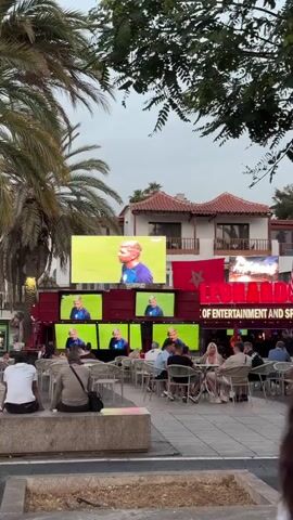 A wide shot captures an outdoor seating area with people watching multiple screens displaying a soccer match. The screens are stacked in two rows, with a large central screen and smaller screens below. Palm trees frame the scene, and a building with a red awning and text reading "LEOF ENTERTAINMENT AND S." is visible in the background. The sky is overcast.
