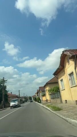 The video shows a street view from a moving vehicle. The sky is blue with scattered white clouds. On both sides of the asphalt road are houses with red tile roofs and light-colored walls, typical of a residential area. Some houses have fences and small trees or bushes in front of them. There are utility poles with wires running along the street. Several cars are visible on the road, including a white van on the left and other passenger cars further ahead. The houses are of varying architectural styles, suggesting a developed but possibly older neighborhood. The road has white lane markings. The overall impression is a sunny day in a European village or town.