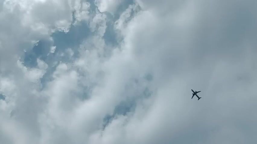 A lone airplane is captured in silhouette against a backdrop of a cloudy sky. The aircraft appears to be a commercial jetliner with its wings and fuselage clearly visible. The clouds are a mixture of white and gray, suggesting a partly cloudy or overcast day. The airplane is in the upper right quadrant of the frame and moves steadily across the sky. The perspective is from below, looking up at the aircraft. No other objects or people are visible in the frame. The lighting is somewhat diffused, indicating that the sun may be behind the clouds. The overall scene is one of aerial transit against a natural sky formation.
