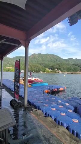 The video shows a body of water with a floating dock. The dock is made of blue modular plastic pieces with orange accents. On the dock, there are two inflatable structures, one blue and one red, and a small red and white boat. In the background, there are green, tree-covered hills and a bright blue sky with scattered clouds. A section of a covered structure with a red roof is visible in the foreground, casting shadows on the wet surface of what appears to be a walkway. A sign with text in a foreign language is placed on the left side of the dock. The camera pans slightly, revealing more of the water and the surrounding landscape.